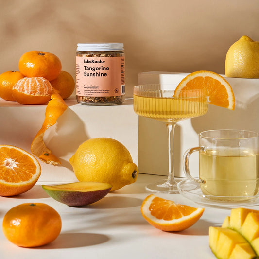 A jar of Lake and Oak 'Tangerine Sunshine' organic tea blend surrounded by various citrus fruits and two glasses of brewed tea.