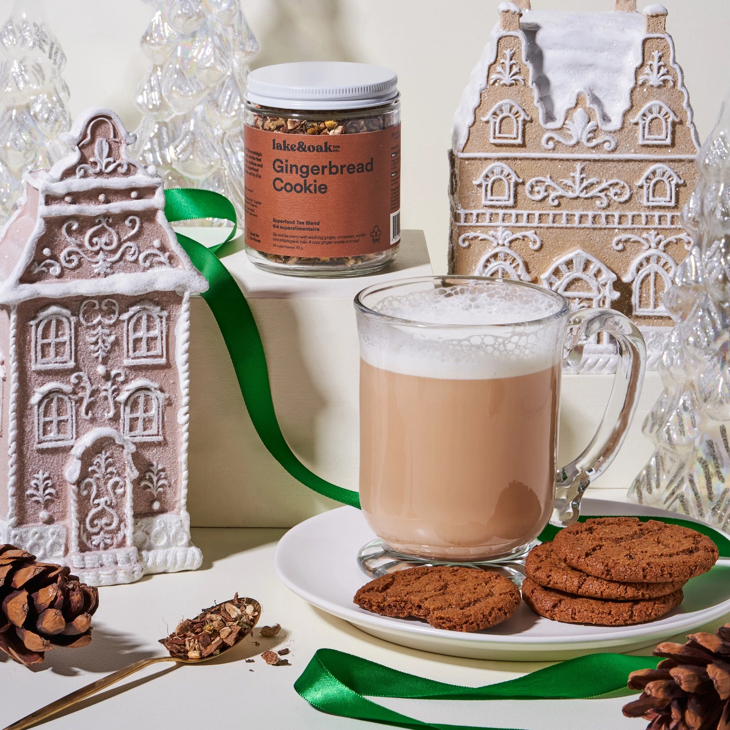 A glass jar of Lake and Oak 'Gingerbread Cookie' organic ginger tea blend in the background, surrounded by artificial gingerbread houses and assorted christmas decorations next to a brewed gingerbread cookie tea latte in the foreground. There are gingerbread cookies next to the tea latte.