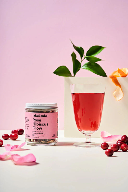 Jar of 'Rose Hibiscus Glow' with a glass of pink drink and cranberries on a light pink background