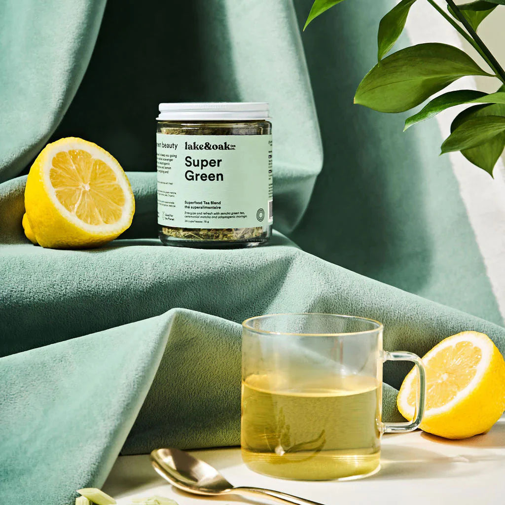 A jar of Lake and Oak Super Green tea blend positioned on a flowy green fabric with sliced citrus around it and a brewed cup of green tea in the foreground.