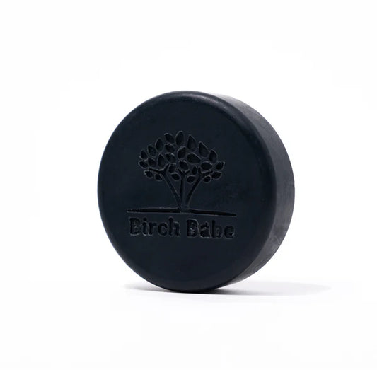A round charcoal facial cleansing bar with the Birch Babe brand logo embossed on it.