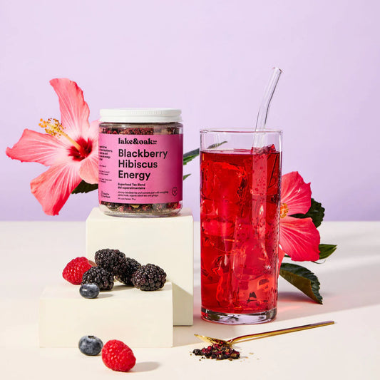 Jar of Lake and Oak 'Blackberry Hibiscus Energy' organic tea blend with a glass of red drink and flowers on a light purple background.